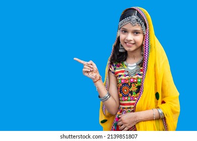 Happy little indian girl kid wearing traditional colorful rajasthani outfit pointing finger over blue background. Advertising Your Text. Look There, Advertisement Banner. blank space.