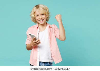 Happy little kid boy 10s wearing pink shirt using mobile cell phone typing sms message doing winner gesture isolated on blue turquoise background children studio portrait. Childhood lifestyle concept