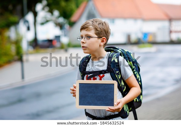 Happy little\
kid boy with glasses and backpack or satchel. Schoolkid on the way\
to middle or high school. Child outdoors on the street. Back to\
school. Kid holding empty chalk\
desk
