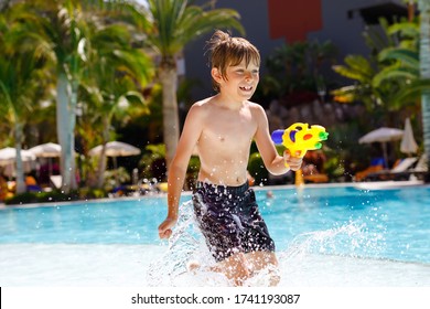 Happy Little Kid Boy Jumping In The Pool And Having Fun On Family Vacations In A Hotel Resort. Healthy Child Playing In Water With A Water Gun. Laughing Running Child Splashing