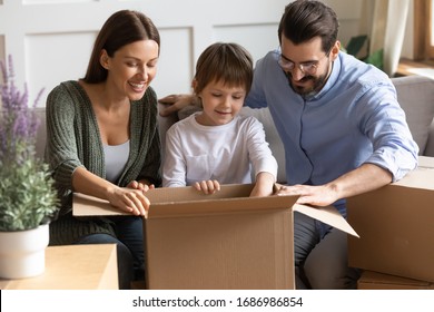 Happy Little Kid Boy Sitting On Sofa Between Smiling Parents, Unboxing Family Belongings Together After Moving In New Apartment. Joyful Family Of Three Unpacking Cardboard Boxes With Stuff At Home.