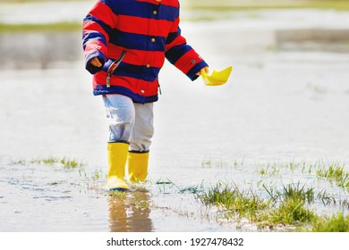 Happy Little Kid Boy In Yellow Rain Boots Playing With Paper Ship Boat By Huge Puddle On Spring Or Autumn Day. Active Leisure For Children. Funny Child Having Fun Outdoors, Wearing Colorful Clothes.