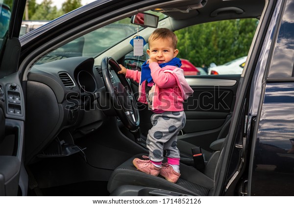 Happy Little kid in a car.\
Stands in the driver\'s seat and holds the wheel. The car door is\
open. Infant driving. Baby looks thoughtfully out of the\
car.
