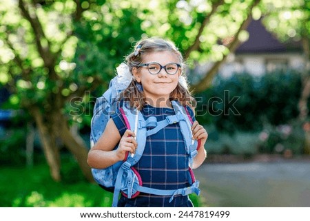 Happy little kid girl with eye glasses with backpack or satchel and big school bag on the first day of school. Healthy adorable child outdoors. Back to school