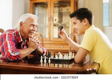 Happy Little Kid Playing Chess With Senior Man At Home. Family Relationship With Grandfather And Grandson. Grandpa And Male Grandchild Playing Board Game