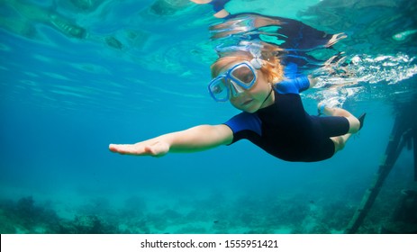 Happy Little Kid In Snorkeling Mask And Wetsuit Jump And Dive Underwater In Coral Reef Sea Lagoon. Family Travel Lifestyle In Summer Adventure Camp. Swimming Activities On Beach Vacation With Child.