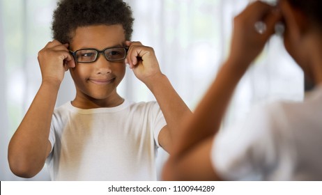 Happy little kid trying on eyeglasses, looking in mirror satisfied good eyesight