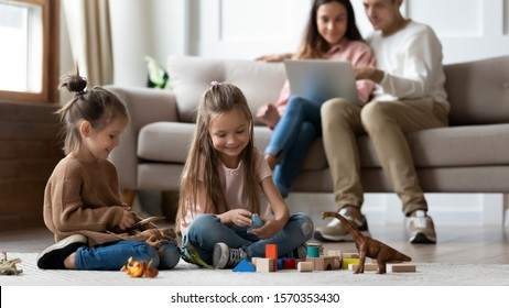 Happy little kids siblings preschool daughters having fun playing toys on floor while parents sit on sofa using laptop in comfy living room, family relaxing at home enjoying cozy leisure lifestyle