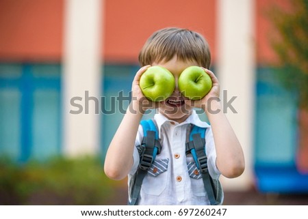 Happy little preschool kid boy with two green apples like his eyes. Boy behind shoulders have backpack. First day to school or nursery. Warm day in an early autumn. Back to school concept.