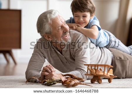 Happy little schoolboy lying on older senior grandfather on floor carpet, having fun together in living room. Joyful different generations family playing, enjoying free weekend time at home.