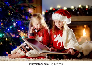 Happy little sisters reading a story book together by a fireplace in a cozy dark living room on Christmas eve. Celebrating Xmas at home.