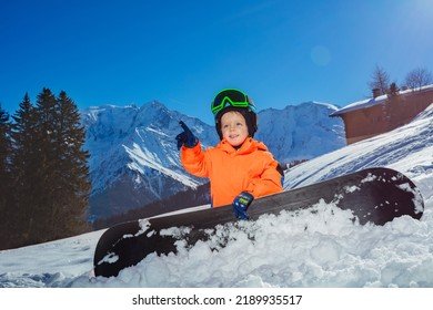 Happy little snowboarder boy point finger and smile on alpine ski track sit wearing helmet ang mask over high mountain
