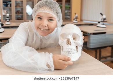 A happy little teenage girl holds an anatomical model of a human skull in her hands. Schoolgirl in a white coat at a science lesson. High quality photo - Powered by Shutterstock