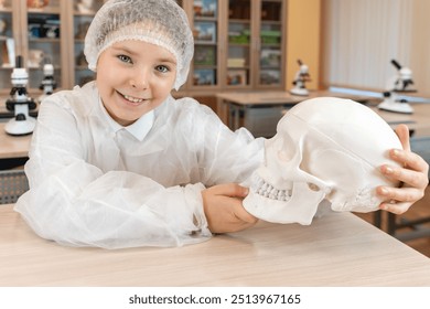 A happy little teenage girl holds an anatomical model of a human skull in her hands. Schoolgirl in a white coat at a science lesson. High quality photo - Powered by Shutterstock