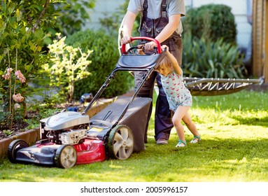 Happy Little Toddler Girl And Middle-aged Father With Lawn Mower. Family, Daughter, Preschool Child And Dad Cut The Lawn. Portrait Of Family Working In Garden, Trimming Grass. Garden Works In Summer.