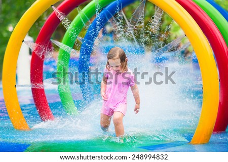 Happy little toddler girl running through a fountain having fun with water splashes in a swimming pool enjoying day trip to an aqua amusement park during summer family vacation