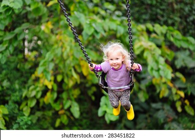 Happy little toddler girl wearing gumboots and warm vest having fun on a swing in the park on a chilly autumn or summer day