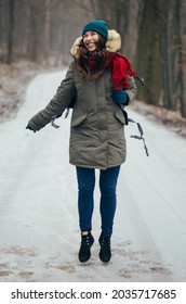 Happy lonely attractive young girl in warm clothing with backpack jumping in forest or park. Single woman loves to walk alone on snow covered road through alley of trees. Hiking, enjoying fresh air