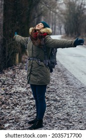 Happy lonely attractive young girl in warm clothing with backpack standing in forest or park. Single woman loves to walk alone on snow covered road through alley of trees. Hiking, enjoying fresh air