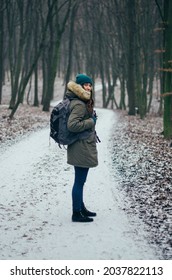 Happy lonely attractive young girl in warm clothing with backpack standing in forest or park. Single woman loves to walk alone on snow covered trail through alley of trees. Enjoying fresh air. Hiking
