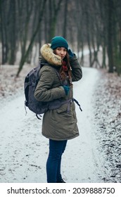Happy lonely attractive young girl in warm clothing with backpack standing in forest or park. Single woman loves to walk alone on snow covered trail through alley of trees. Hiking, enjoying fresh air