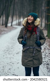 Happy lonely attractive young girl in warm clothing with backpack standing in forest or park. Single woman loves to walk alone on snow covered trail through alley of trees. Hiking, enjoying fresh air