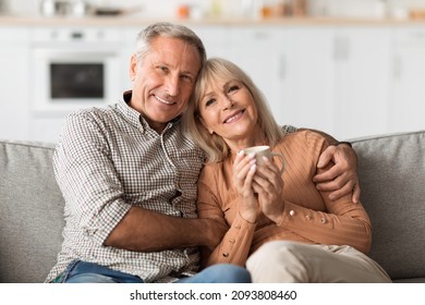 Happy Long-Lasting Marriage. Older Husband Hugging Wife While She Holding Cup Of Coffee, Posing Smiling To Camera Sitting On Couch At Home. Retirement Lifestyle And Happiness - Powered by Shutterstock