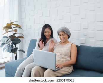 Happy Lovely Asian Senior Woman, Mother White Hair And Beautiful Young Female, Daughter Waving Greeting At Laptop Computer Screen While Sit On Couch In White Room. Meet Family By Video Call At Home.