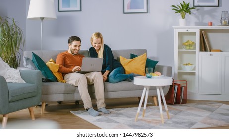 Happy Lovely Couple Sits On A Sofa At Home, Boyfriend Holds Laptop On Knees, Browsing Through Internet, Doing E-Shopping, Using Streaming Services. Happy Family In Cozy Living Room.