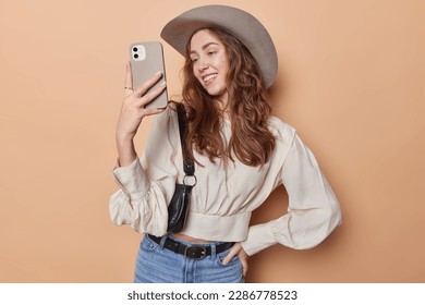 Happy lovely woman with dark wavy hair makes sefie photo on smartphone smiles happily dressed in stylish outfit isolated over brown background. Brunette female model in cowboy hat has video call