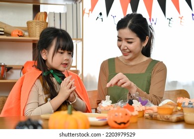 Happy And Lovely Young Asian Girl In Halloween Costume Enjoys Decorating Cupcakes With Her Mom At Home. Happy Halloween Holiday Concept