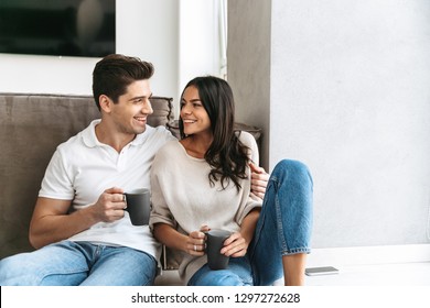 Happy lovely young couple holding cups while sitting on a floor at the couch at home - Shutterstock ID 1297272628
