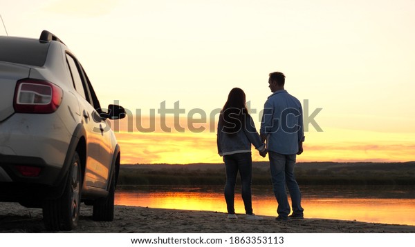 happy lovers travelers man and woman stand next to\
car and admire beautiful sunset on beach. tourists travel by car,\
hug, admiring sunrise, river. Free travelers, tourists. family\
travel by car.
