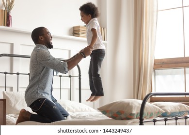 Happy Loving African American Young Dad Enjoy Playing With Toddler Son Have Fun Together In Bedroom, Smiling Caring Black Father Hold Lift Laughing Small Boy Child Jumping On Bed Spend Weekend At Home