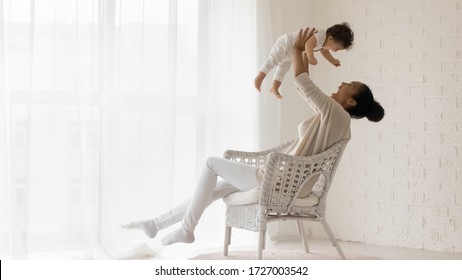 Happy Loving African American Young Mother Sit In Chair At Home Relax Play With Funny Little Newborn Baby Girl, Smiling Biracial Mom Have Fun Lift In Air Enjoy Family Weekend With Cute Infant Child