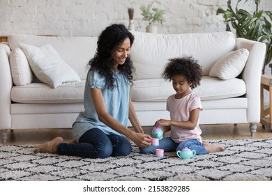 Happy Loving African Mom And Sweet Preschool Daughter Kid Pretending Drinking Coffee In Earning Game, Playing Tea Party With Toy Cups, Teapot, Dish On Heating Carpeted Floor In Living Room