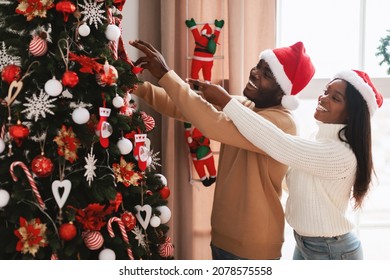 Happy Loving Black Family Decorating Beautiful Christmas Tree Together In Living Room. Excited Lady And Guy In Red Santa Claus Hat Preparing For Xmas Holiday Celebration Making House Cozy And Festive