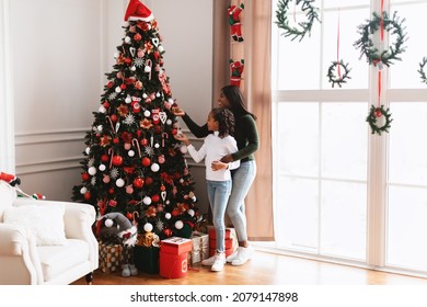 Happy Loving Black Family Decorating Beautiful Christmas Tree Together In Living Room. Excited African American Lady And Girl Preparing For Xmas Holiday Celebration Making Their House Cozy And Festive