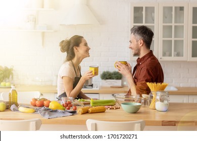 Happy Loving Couple Chatting During Coffee Break Standing At Home Kitchen. Handsome Husband And Beautiful Wife Together. Woman In Apron Cook Fresh Vegetable Salad. Man Entertain Beloved Soulmate