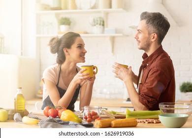 Happy Loving Couple Drinking Tea Or Coffee On Kitchen. Cheerful Smiling Young Man And Woman Rest Having Break During Cooking Process At Home. Married Girl And Guy Talking, Sharing Nice Emotions