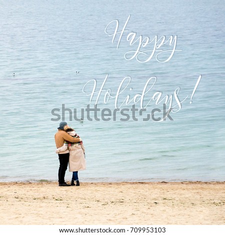 Happy loving couple embracing wearing warm clothes standing on ocean coast. Cold season, sea shore, empty beach in autumn or winter. Happy holidays inscription, greeting card.