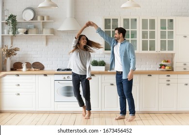 Happy loving couple having fun in kitchen, dancing together, celebrating relocation or anniversary, handsome young husband holding beautiful wife hand, moving to favorite music, enjoying date