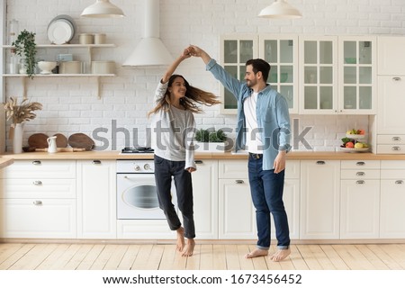 Happy loving couple having fun in kitchen, dancing together, celebrating relocation or anniversary, handsome young husband holding beautiful wife hand, moving to favorite music, enjoying date