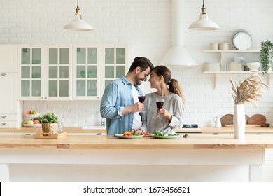 Happy loving couple holding wine glasses, touching foreheads, enjoying tender moment, romantic date, standing in modern kitchen at home together, smiling wife and husband celebrating anniversary