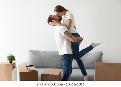 Happy loving couple kissing feeling excited about moving into new home, man lifting holding woman in hands standing among cardboard boxes, glad tenants enjoying relocation, starting living together