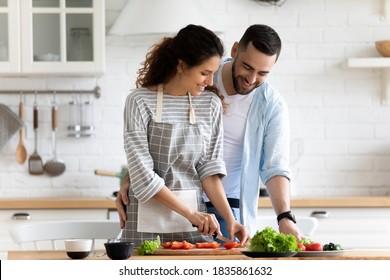 Happy loving couple preparing dinner in modern kitchen together, hugging, standing at table, smiling young woman wearing apron cutting vegetables for salad, spouses enjoying leisure time at home - Powered by Shutterstock