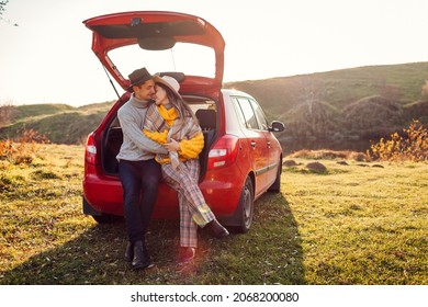 Happy loving couple relaxing in car trunk by autumn lake hugging. Man and woman travel by auto. Fall season. Young people wear warm clothes