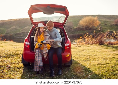 Happy loving couple relaxing in car trunk by autumn pond drinking tea from vacuum flask. Man and woman travel by auto. Young people enjoy fall trip