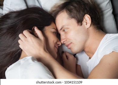 Happy loving couple relaxing on bed at home, young man touching smiling beautiful woman face, looking in the eyes stroking caressing, enjoying togetherness and tenderness in love, top close up view