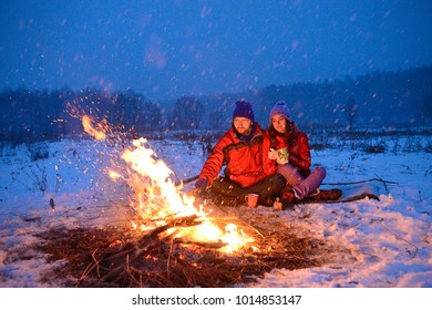 Happy Loving Couple Sitting At A Fire In The Snow Among The Fields In The Winter Against The Background Of The Forest And The Sky On Valentine's Day.